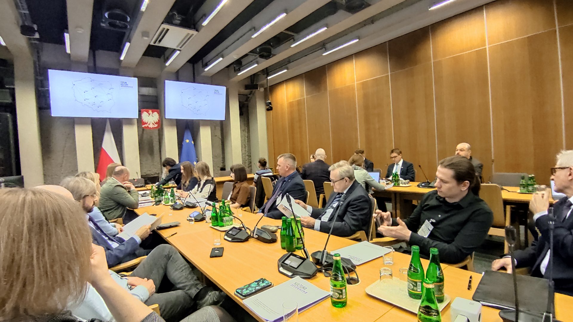 Na zdjęciu widać grupę osób siedzących przy stołach, wyglądają na obradujących podczas posiedzenia jakiegoś większego zespołu.