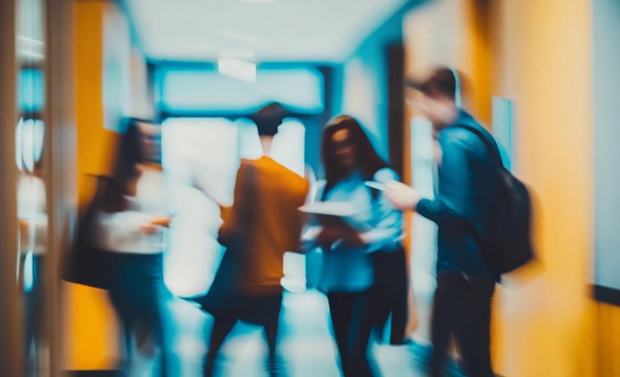 Grafika przedstawia rozmytą scenę w korytarzu lub holu – widać kilka postaci w ruchu (kobietę, mężczyzn), jedna osoba trzyma jakieś dokumenty. Tło jasne, kolory stonowane z akcentem ciepłych odcieni (np. światło żółtawe / pomarańczowe), ogólne wrażenie pośpiechu i nieostrości.