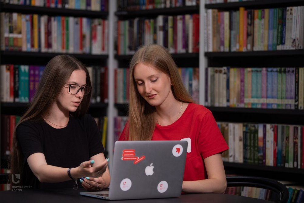 Dwie studentki siedzą przy laptopie w bibliotece i rozmawiają. 