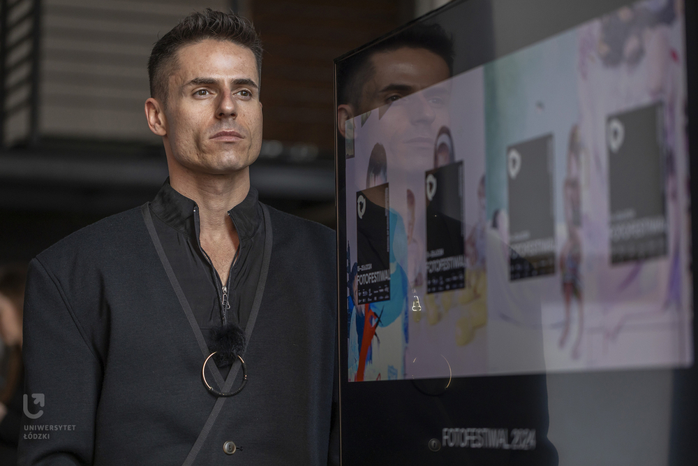 na zdjęciu Bartosz Kałużny, brunet stojący obok ekranu na zdjęciu Bartosz Kałużny, brunet stojący obok ekranu