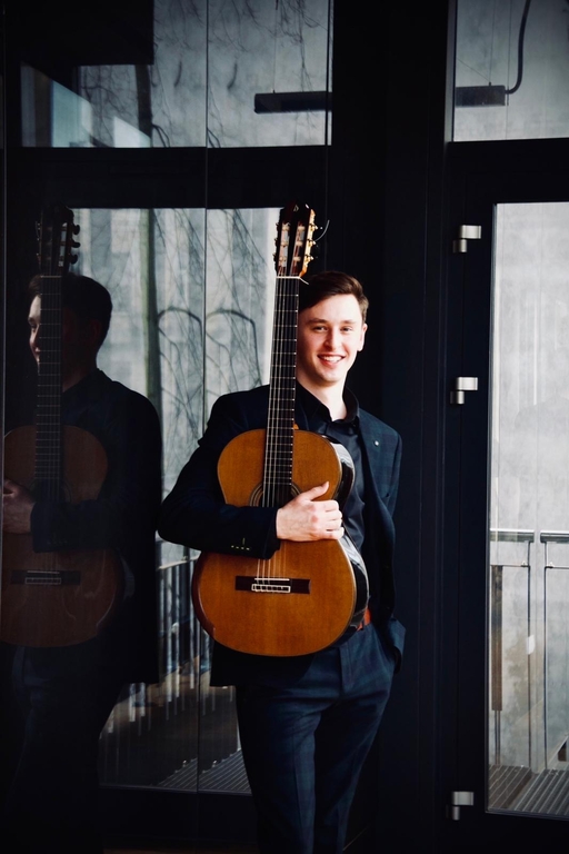 Maksym Niewczas ubrany w ciemny garnitur, trzymający gitarę
