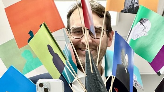 Na zdjęciu widać mężczyznę, który trzyma chorągiewki zatknięte w stożku ze styropianu. Mężczyzna trzyma w drugiej dłoni smartfon i robi nim zdjęcie.