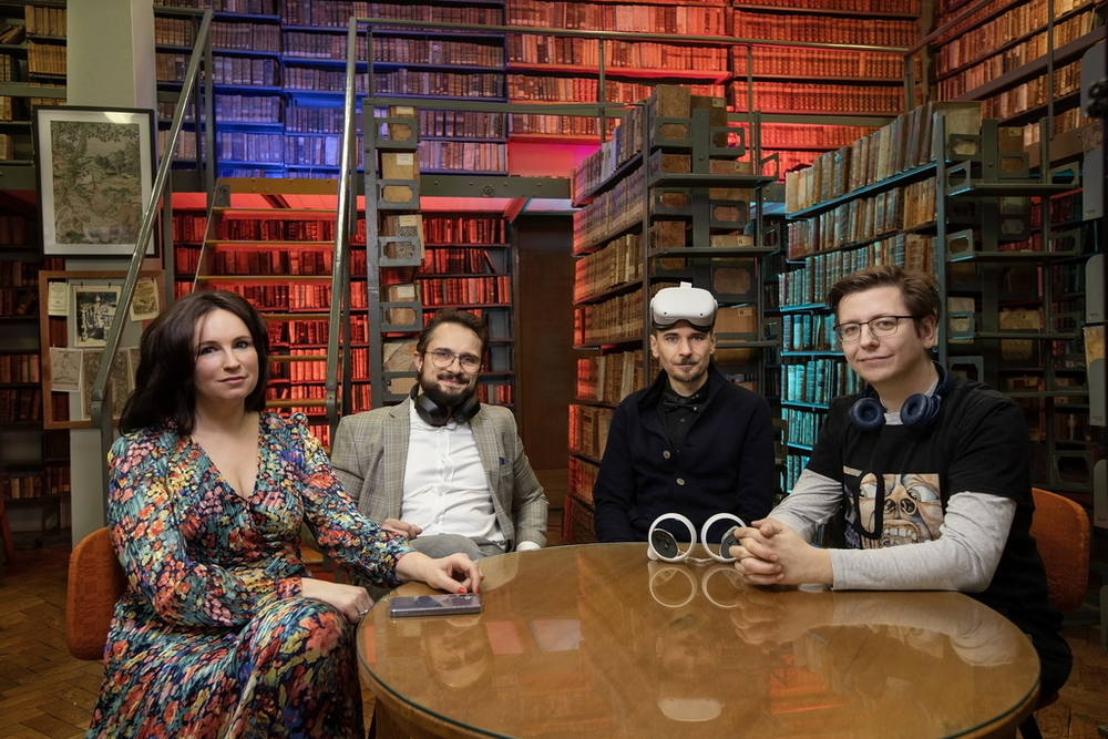 Cztery osoby siedzące przy stole, patrzą w stronę obiektywu. Otaczają je regały książek. Pomieszczenie wypełnia kolorowa poświata. Każda z osób ma przy sobie jakieś urządzenie elektroniczne: słuchawki, telefon lub okulary do projekcji wirtualnej.