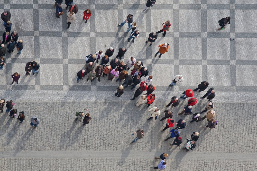 crowd of people in center of town, top view Na zdjęciu widać grupę ludzi. Widziani są z góry, z lotu ptaka. Stoją w kilku miejscach na chodniku lub kamiennych płytach.