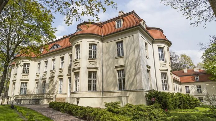 Na zdjęciu widoczny jest duży budynek, który wygląda jak pałac. Obiekt stoi wśród drzew. Widać, że jest słoneczny dzień.