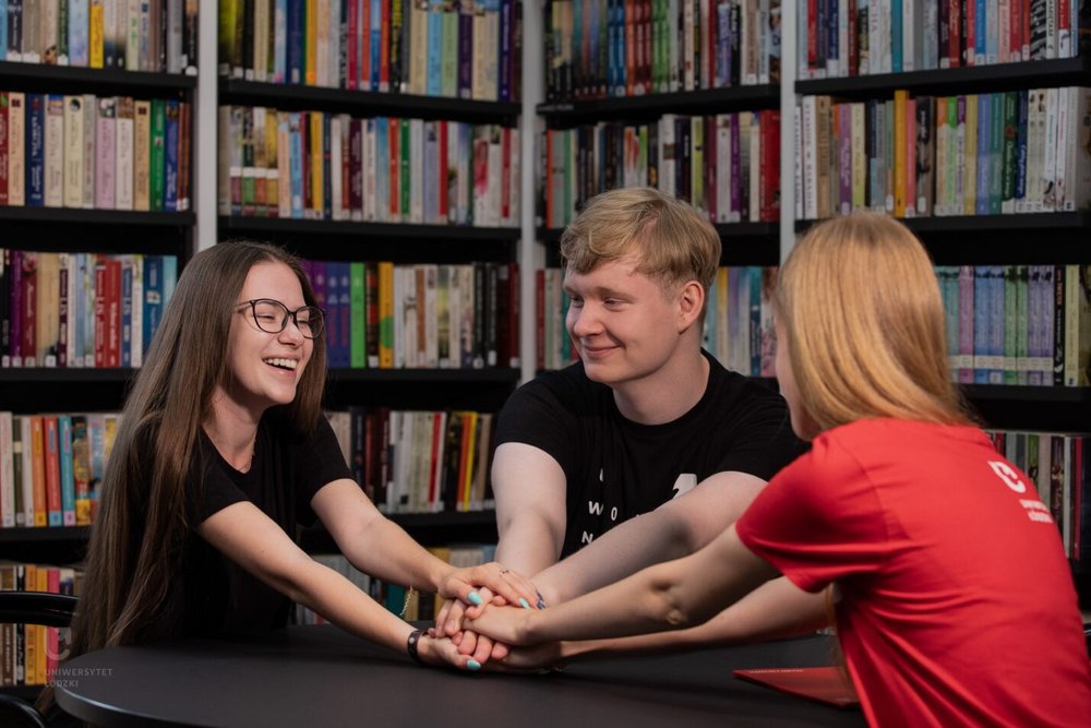 2 studentki i student siedzą w bibliotece przy regałach z książkami i trzymają się za ręce 