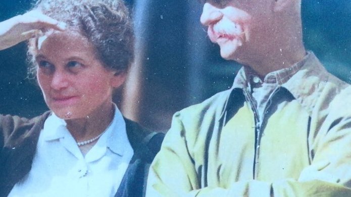 Na zdjęciu widać mężczyznę i kobietę stojących i patrzących przed siebie. Kobieta trzyma prawą dłoń nad czołem i przysłania oczy przed ostrym słońcem, którego blask widoczny jest na zdjęciu.