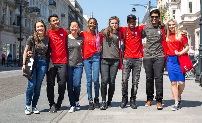 Grupa młodych ludzi różnych kolorów skóry stoi na ulicy Piotrkowskiej w Łodzi. Osoby na zdjęciu uśmiechają się, mają na sobie koszulki UŁ.