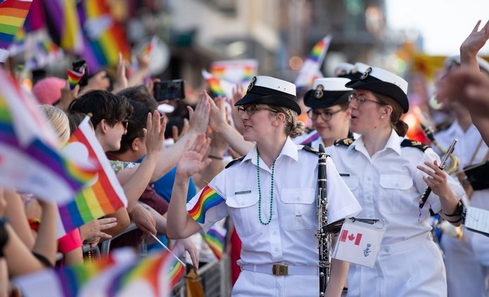 The photo shows a crowd of people, a procession. In the foreground, women in white uniforms can be seen smiling and giving high-fives to people standing on the left side of the frame. In the background, a large number of colorful rainbow flags can be seen