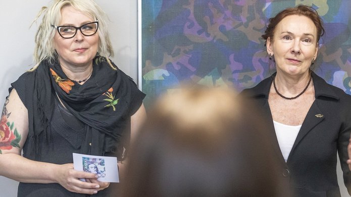 Na zdjęciu widoczne są dwie kobiety. Stoją twarzami do nas. Za nimi widczone są wiszące na ścianie obrazy. Kobiety widzimy od pasa w górę. 