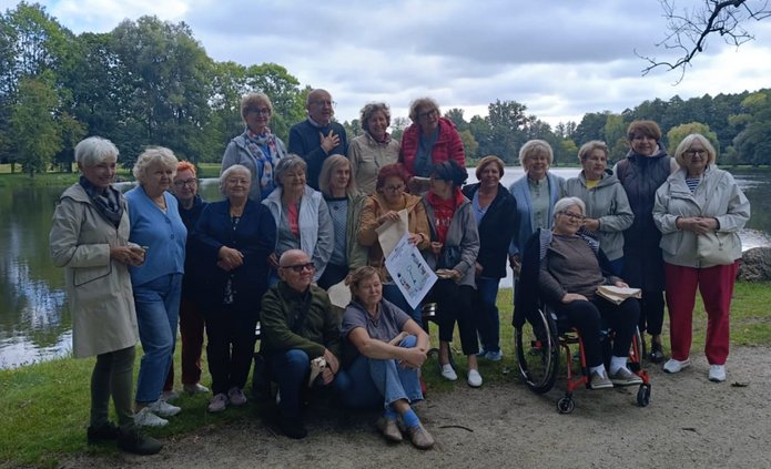 Na zdjęciu widać grupę około 20 osób. To mężczyźni i kobiety w dojrzałym wieku, pozują do zdjęcia grupowego. Stoją w plenerze. Jest to pora letnia.