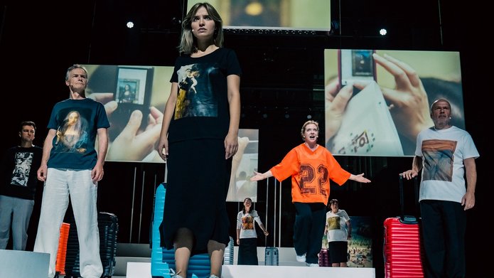 Na zdjęciu widać grupę osób stojących i patrzących w jeden wspólny punkt. To kobiety i mężczyźni ubrani jak turyści, koszulki sportowe spodnie i obuwie. Przy każdej osobie stoi walizka. W tle widoczne na wielkich ekranach zdjęcia z podróży, z turystami.