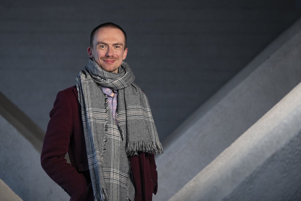 Zdjęcie dr inżyniera Błażeja Ciarkowskiego - krótkowłosy, szczupły blondyn, ubrany w szary komplet z szalem. 