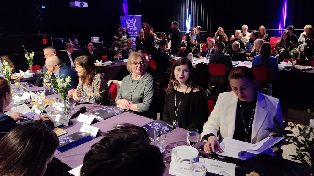 Na zdjęciu widoczne są osoby siedzące za stołem. Jest to spora grupa. Osoby debatują w sali, która wygląda jak sala teatralna. Jest dość ciemno, a oświetlenie jest teatralne, z fioletowym odcieniem. 