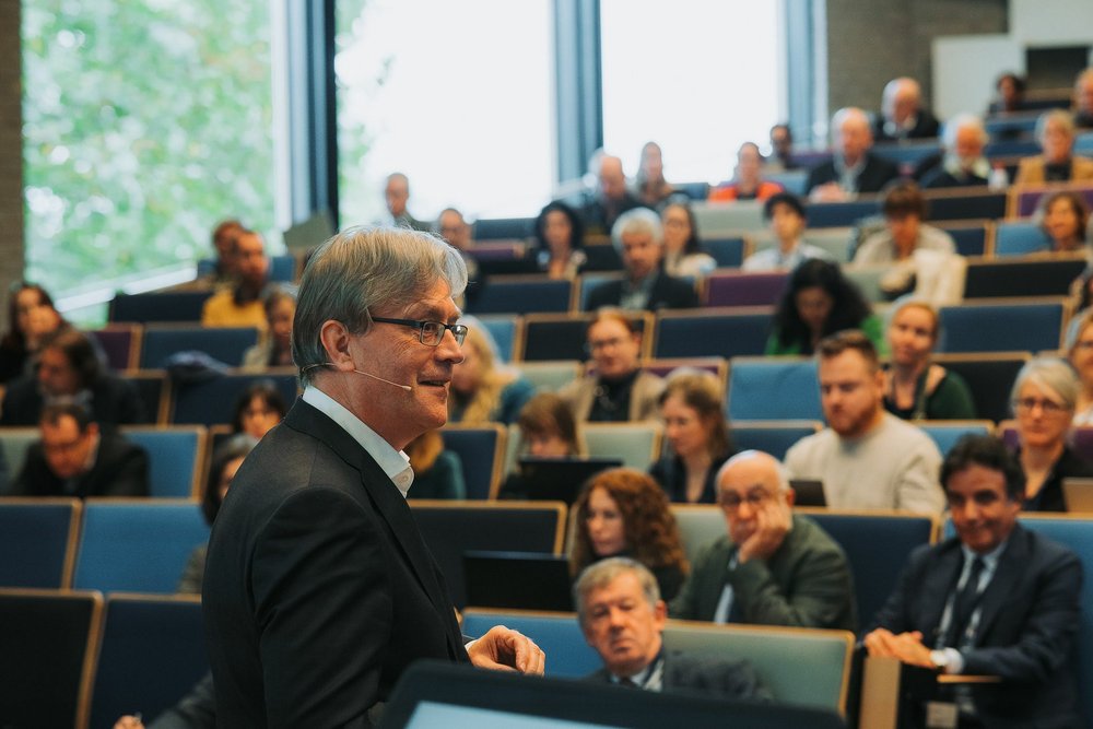 mężczyzna patrzy w prawo i mówi do mikrofon; w tle publiczność na auli uniwersyteckiej