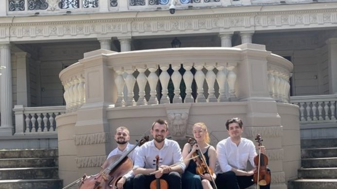 Kobieta i 3 mężczyzn - członkowie zespołu Vivenda String Quartet, siedzą z instrumentami smyczkowymi przed budynkiem