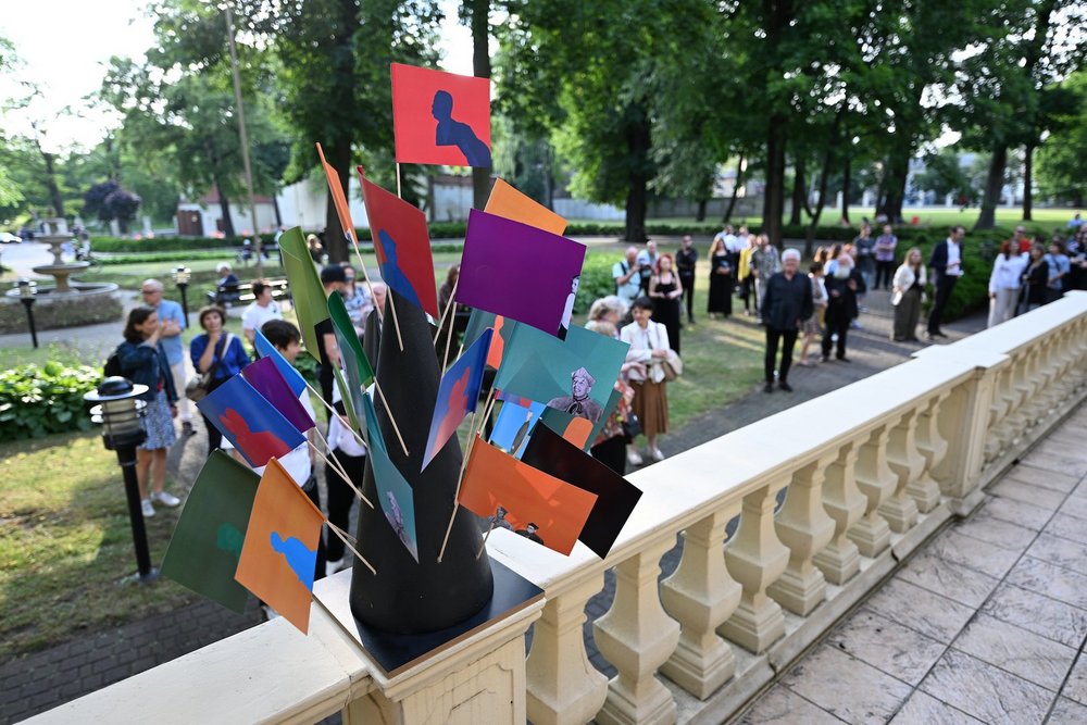 Na zdjęciu widać park, w którym jest dużo ludzi. Jest wiosna, drzewa są zielone. Na pierwszym planie widoczna balustrada z betonu na której stoi stojak z kolorowymi chorągiewkami. W tle chodzą wspomniani ludzie.