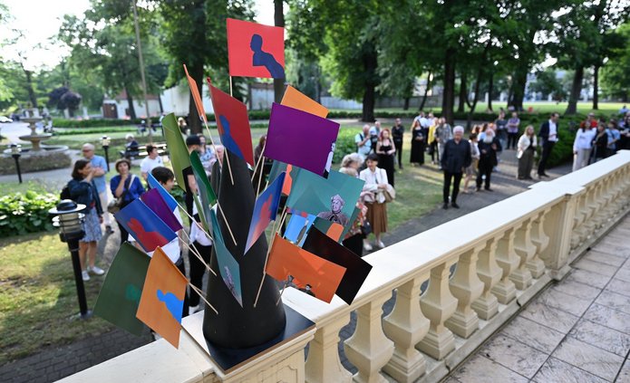 The photo shows a park crowded with people. It's spring, and the trees are green. In the foreground, a concrete railing is visible, with a stand of colorful flags on it. In the background, the aforementioned people are walking.