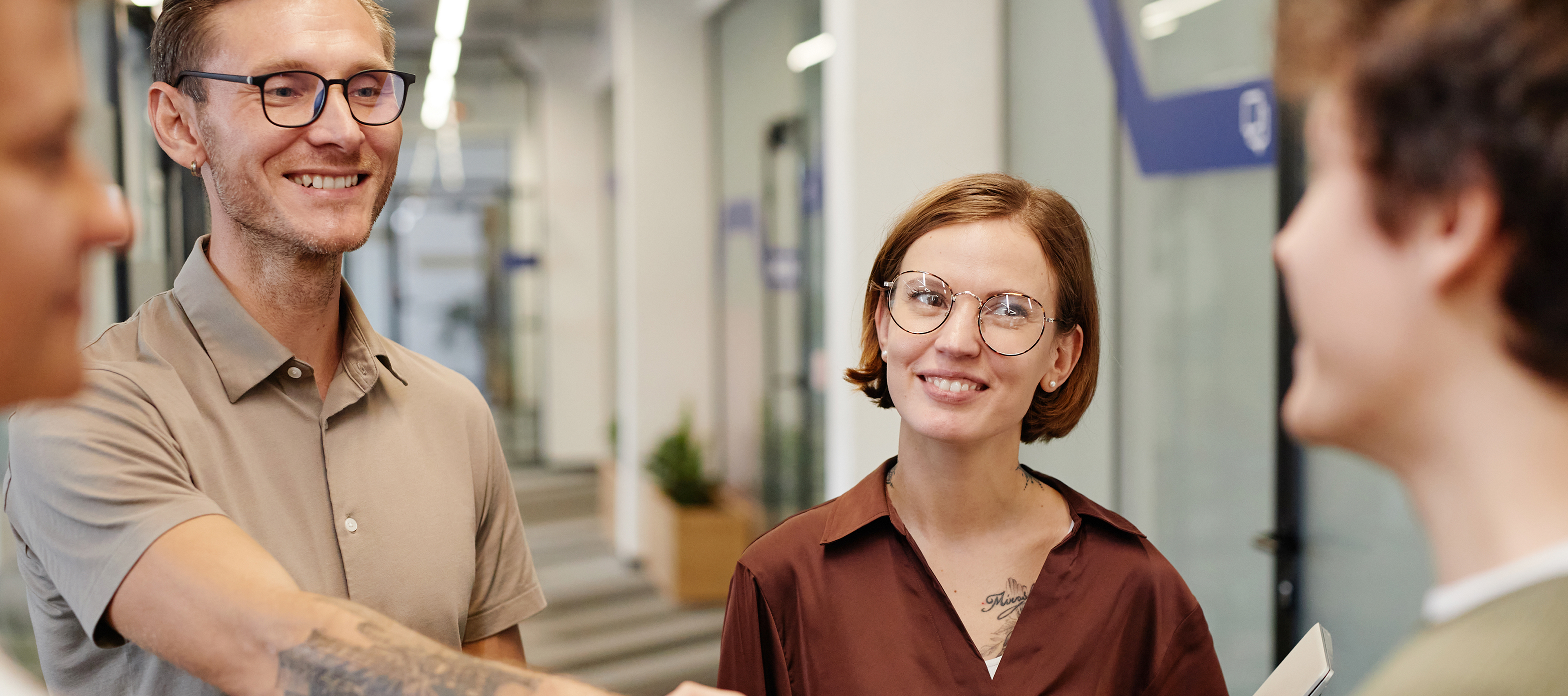 kolorowe zdjęcie przedstawiające cztery młode osoby (dwóch mężczyzn i dwie kobiety) stojących w kółku podczas rozmowy; uśmiechnięty mężczyzna w okularach stojący na pierwszym planie wita jedną z tych osób