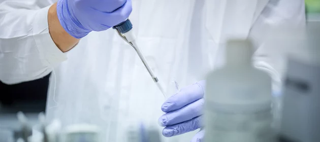 Pipeta trzymana przez osobę w białym fartuchu i gumowych rękawiczkach w laboratorium