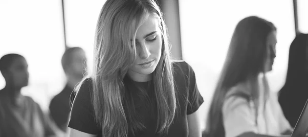 Młoda kobieta siedząca w sali. W tle widać inne osoby siedzące w sali.